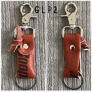 以下が含まれることがあります： 赤色の革製ホルダーと銀色の留め金が付いた2つのキーホルダー。ホルダーには鍵用のループがあり、「GOOD」の文字が刻印されています。留め金は金属リングに取り付けられています。