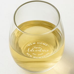 May include: A stemless wine glass filled with a light yellow liquid. The glass has white text that reads "NEW HOME, NEW ADVENTURE, NEW MEMORIES, EST. 2025" in a circular design. The glass is on a white background.
