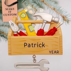 May include: A wooden toolbox Christmas ornament with a hammer, screwdriver, pliers, and level. The toolbox is yellow and brown with the name "Patrick" and the word "YEAR" on it. A silver wrench hangs from the bottom of the ornament.