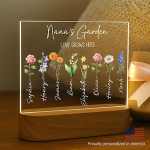 May include: A personalized acrylic light-up sign with a wooden base. The sign features a floral design with the text "Nana's Garden, Love Grows Here" and a list of names: Sophia, Henry, James, Liam, Elizabeth, Olivia, Hailey, and Noah.