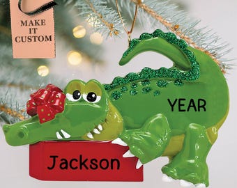 Può includere: Addobbo natalizio a forma di alligatore verde che tiene una scatola regalo rossa con un fiocco rosso. L'addobbo ha il nome "Jackson" e la parola "YEAR" su di esso.