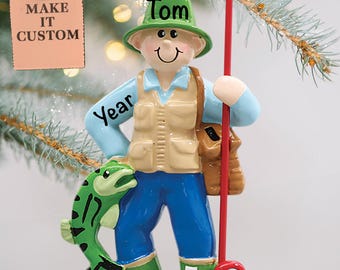 May include: A personalised Christmas bauble featuring a cartoon fisherman wearing a green hat, blue trousers, and green boots. The bauble has the name "Tom" written on the hat and the word "Year" written on the fisherman's jacket. The fisherman is holding a fishing rod and a green fish.