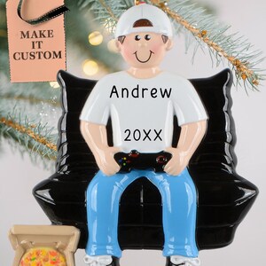 May include: A personalized Christmas ornament featuring a cartoon figure of a person wearing a white cap, white shirt with the name "Andrew" and the year "20XX", blue jeans, and black and white sneakers. The figure is holding a game controller and sitting on a black bench. A pizza box is at the bottom.