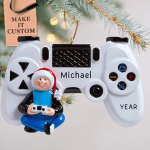 May include: White and black video game controller Christmas ornament with a cartoon figure of a person wearing a Santa hat and holding a blue video game controller. The ornament is personalized with the name "Michael" and the year.