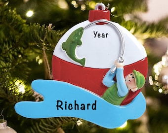 May include: A blue and red Christmas bauble shaped like a fishing bobber with a cartoon fisherman holding a fishing rod. The bauble has the text "Year" and "Richard" on it.