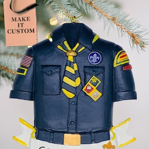 May include: A navy blue Boy Scout uniform ornament with a yellow and black striped tie. The ornament has a banner that reads "Connor YEAR". The tag reads "MAKE IT CUSTOM".