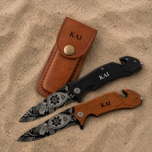 May include: A tan leather sheath with a brass snap button and the text KAI, alongside two folding knives with black blades featuring white floral patterns. One knife has a black handle and the other a wood handle, both marked with the text KAI.