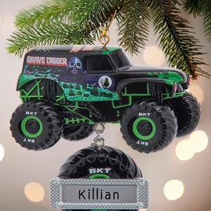 May include: Black and green Grave Digger monster truck ornament with the name "Killian" on a silver tag. The truck has large black tires with green accents and the text "BKT". The ornament hangs from a gold-colored hook.