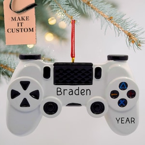 May include: White plastic video game controller Christmas ornament with black buttons and a black and white textured top. The ornament has the name "Braden" printed on the top and the word "YEAR" printed on the bottom.