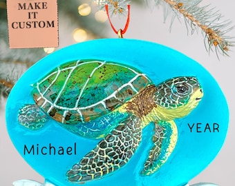 May include: A personalised Christmas ornament with a sea turtle design. The turtle is green, brown, and yellow, set against a blue background. The ornament includes the name "Michael" and "Grand Bahama". The tag says "MAKE IT CUSTOM".
