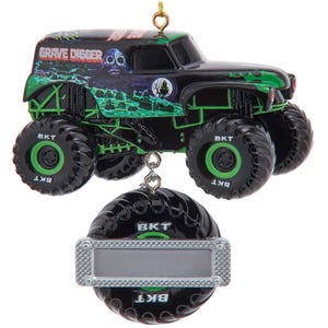 May include: A black and green Grave Digger monster truck ornament with large tires and the text "Grave Digger." The ornament includes a silver tag. The truck has a skull and graveyard design.