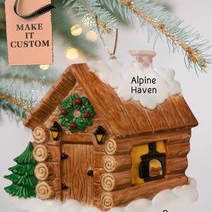 May include: A brown wooden cabin Christmas ornament with a white snowy roof, a green wreath on the door, and a fireplace with flames. The ornament has the text "Alpine Haven" and "YEAR" on it.