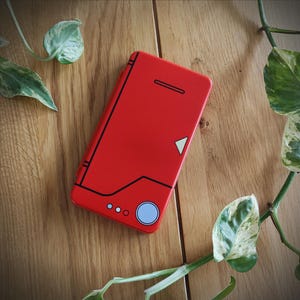 May include: A red phone case with black geometric designs and a light blue circle. The case has a rectangular shape and is lying on a wooden surface. Green and white leaves are in the background.