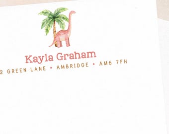 May include: White stationery with a watercolor illustration of a pink dinosaur and palm tree. The name "Kayla Graham" is printed in a reddish-brown color, along with the address "12 Green Lane • Ambridge • AM6 7FH". An envelope is visible.