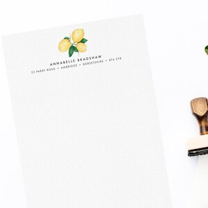May include: A white sheet of paper with a lemon illustration and the text "Annabelle Bradshaw, 23 Parley Road, Ambridge, Berkshire, BT4 5YR". There are three wooden rubber stamps and a white magnolia flower on a white surface.