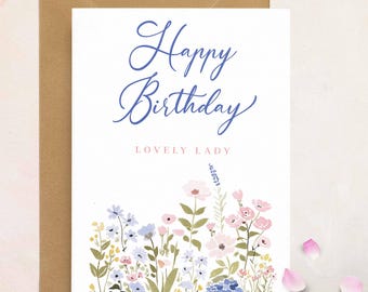 May include: A white greeting card with the words "Happy Birthday" in blue script, and "LOVELY LADY" in pink. The card features a floral illustration at the bottom, with pink, blue, and yellow flowers. The card is inside a brown envelope.