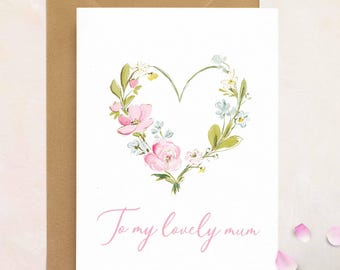 May include: A white greeting card with a floral heart design in pink, green, and blue, with the text "To my lovely mum" in pink script. The card is paired with a brown envelope and scattered pink petals.