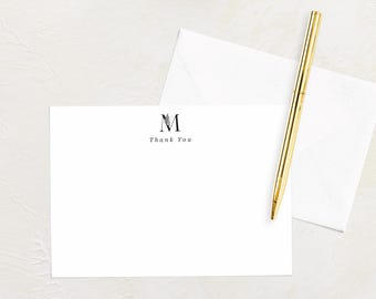 May include: A white stationery set featuring a blank card with the letter "M" and "Thank You" printed in black. A gold pen and a white envelope are also present. The items are arranged on a light-colored surface.
