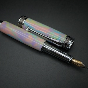 May include: A luxury fountain pen with a pearlescent barrel in pastel colors of pink, blue, and yellow. The pen has silver-colored metal accents and a gold-colored nib. The cap is off, revealing the nib and the pen's design.