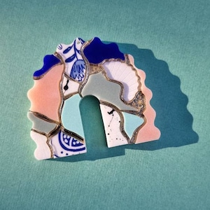May include: A colourful, abstract arch-shaped resin piece with a blue, green, pink, and white mosaic design. The piece has a gold glitter accent.