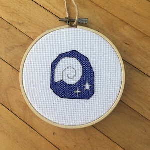 May include: A white embroidery hoop with a blue and white cross-stitch design of a seashell with two stars. The hoop is hanging from a brown string.