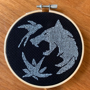 May include: A black embroidery hoop with a white wolf, bird, and plant design. The design is stitched in a cross-stitch pattern.