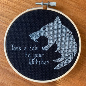 May include: A black and white embroidered hoop art piece with a wolf's head and the text "Toss a coin to your Witcher".