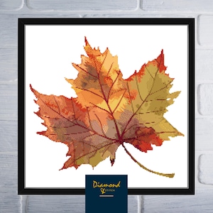 May include: A framed cross-stitch of a fall maple leaf. The leaf is rendered in shades of orange, yellow, and red, with detailed veining. The artwork is set against a white background and framed in black. The lower right corner features a logo that reads "Diamond Stitch".