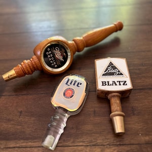 May include: Three beer tap handles. One is a horizontal wooden Miller Genuine Draft handle. Another is a clear acrylic Lite handle with a circular logo. The third is a vertical wooden Blatz handle with a white triangular logo and text.
