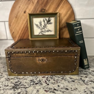 May include: A vintage brass-toned box with studded trim, a small framed botanical print, and a green book titled "A Dictionary of Antique" are displayed on a countertop. A wooden cutting board is in the background.