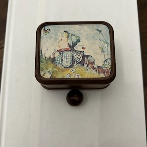 May include: A small, square, brown trinket box with a decorative lid featuring a vintage illustration of a child in a field. The box has a small, round, dark brown knob on the bottom. The box is resting on a white surface.