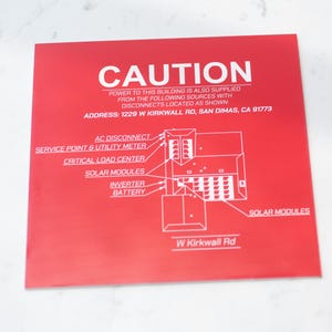 May include: A red square sign with white text and a diagram. The word "CAUTION" is at the top. The sign provides information about power sources and disconnects, including an address: 1229 W Kirkwall Rd, San Dimas, CA 91773.