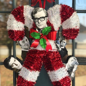 May include: A red and white candy cane shaped Christmas wreath with a holly sprig and a man's face in the center.  Four other men's faces are peeking out from behind the wreath.