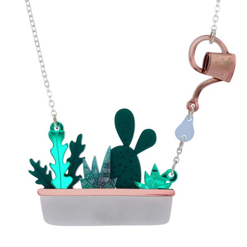 K&ouml;nnte beinhalten: Halskette mit silberner Kette und Anh&auml;nger mit einem Pflanzkasten mit gr&uuml;nen und t&uuml;rkisfarbenen Sukkulenten. Ein Charme in Form einer gie&szlig;kanne in Kupferfarbe ist angebracht. Ein einzigartiges Accessoire.