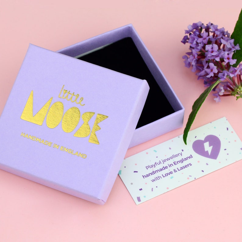 May include: A light purple box with the words "little moose" in gold lettering and the text "handmade in England". The box is open and a small white card with the text "Playful jewellery handmade in England with Love & Lasers" is visible.