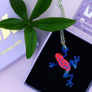 May include: A red and blue frog pendant necklace. The frog is made of acrylic and has a shiny finish. The necklace is silver and has a delicate chain.