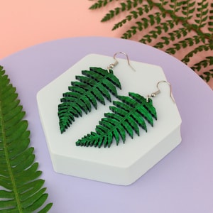 Puede incluir: Un par de pendientes colgantes con forma de hojas de helecho. Los pendientes son de un color verde intenso y vibrante con una apariencia texturizada y en capas que imita la estructura natural de una fronda. Cada pendiente tiene un gancho de metal plateado.