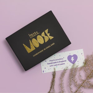K&ouml;nnte beinhalten: Eine schwarze Schachtel mit goldfarbenem Schriftzug "Little Moose Handmade in England" liegt auf einer hellvioletten Oberfl&auml;che. Eine kleine Karte mit Herz- und Blitzgrafik ist ebenfalls zu sehen, zusammen mit dekorativen Zweigen.