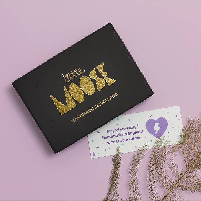 K&ouml;nnte beinhalten: Eine schwarze Schachtel mit goldfarbenem Schriftzug "Little Moose Handmade in England" liegt auf einer hellvioletten Oberfl&auml;che. Eine kleine Karte mit Herz- und Blitzgrafik ist ebenfalls zu sehen, zusammen mit dekorativen Zweigen.
