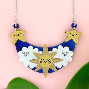 Peut inclure: Un collier avec un pendentif en forme de croissant de lune bleu profond et un soleil doré pailleté au centre. Deux nuages blancs aux visages souriants entourent le soleil, tandis que deux étoiles dorées souriantes ornent les extrémités.