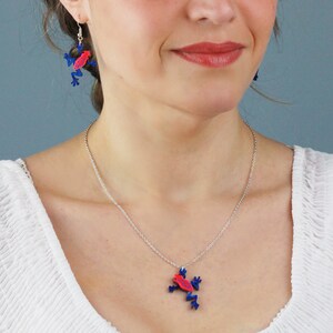 May include: A silver chain necklace with a red and blue frog pendant. The frog is facing to the right and has a detailed design. The necklace is worn by a person with a white shirt.