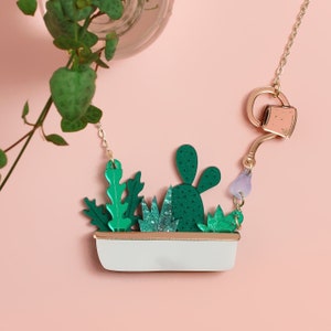 K&ouml;nnte beinhalten: Eine skurrile Halskette mit einer wei&szlig;en Pflanzschale mit gr&uuml;nen Kakteen und Bl&auml;ttern. Eine goldene Kette h&auml;lt den Anh&auml;nger, mit einem Gie&szlig;kannen- und einem lavendelfarbenen Charm. Der Hintergrund ist ein zartes Rosa.