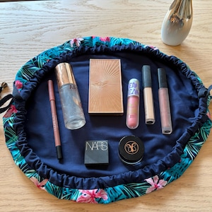 May include: A navy blue drawstring makeup bag with a tropical floral design, containing various cosmetic products. Items include a bronzer, lipsticks, a lip liner, and a setting spray. The bag is open, displaying the contents on a dark blue interior.