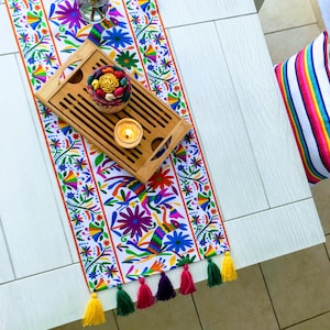 Puede incluir: Un camino de mesa colorido con borlas, con un diseño tradicional mexicano. El camino de mesa está colocado sobre una mesa blanca con una bandeja de madera y una vela en la parte superior.