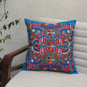 May include: A turquoise blue throw pillow with a colorful floral embroidery design. The pillow is sitting on a white couch with a wooden armrest.