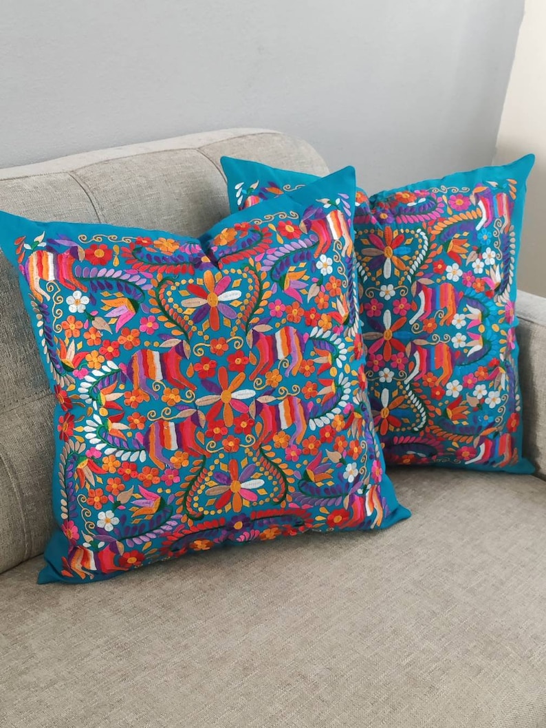 May include: Two turquoise blue throw pillows with colorful embroidered floral patterns. The pillows are on a gray couch.