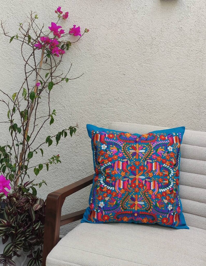 May include: A turquoise blue throw pillow with a colorful floral embroidery design. The pillow is sitting on a white couch with a wooden armrest.