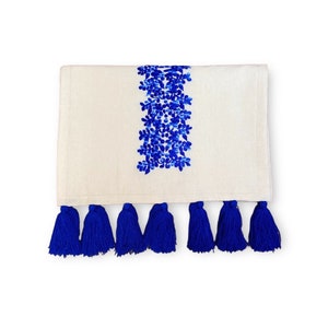 May include: White table runner with a blue floral embroidered design and blue tassels on the bottom edge.