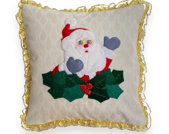 Puede incluir: Una almohada blanca con ribete dorado presenta una imagen bordada de Santa Claus con un gorro rojo y barba blanca, sosteniendo una rama de acebo con bayas rojas.