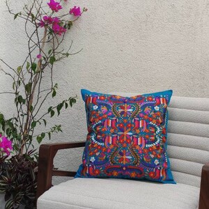 May include: A white chair with a teal blue pillow featuring a colorful floral embroidery pattern. The chair is in front of a white wall with three small decorative houses.